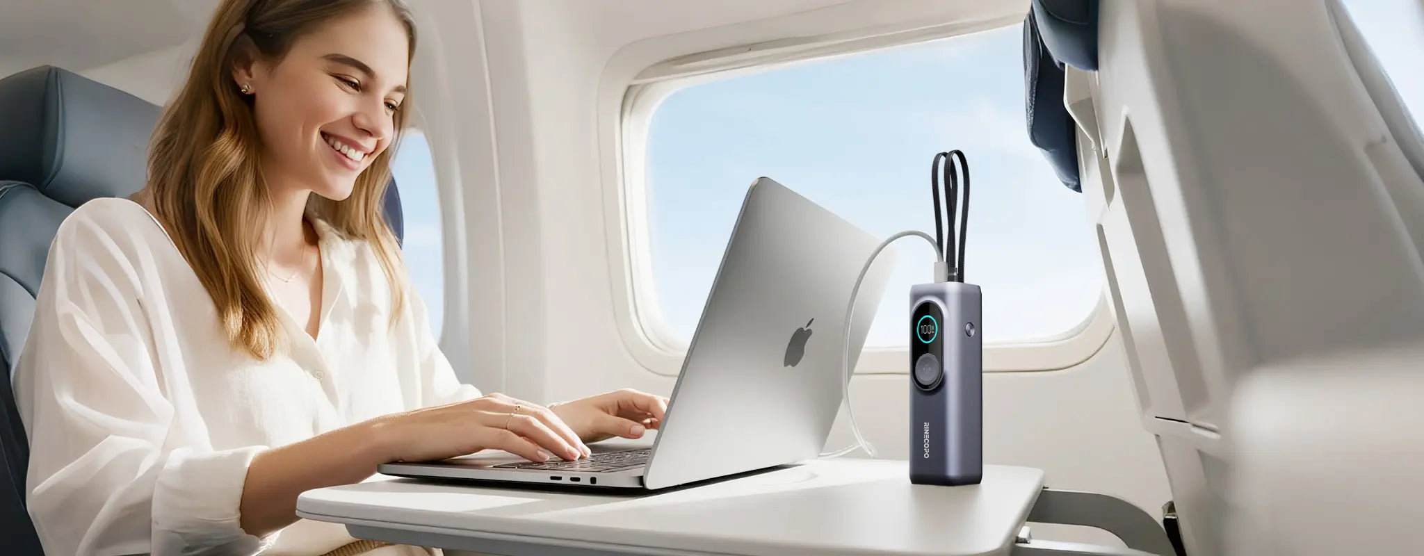 A traveler using the Rinecopo portable power bank to charge a MacBook while on a flight. This high-capacity, airline-approved external battery is the perfect travel companion for long-haul flights, ensuring your business tasks and entertainment stay powered at 30,000 feet